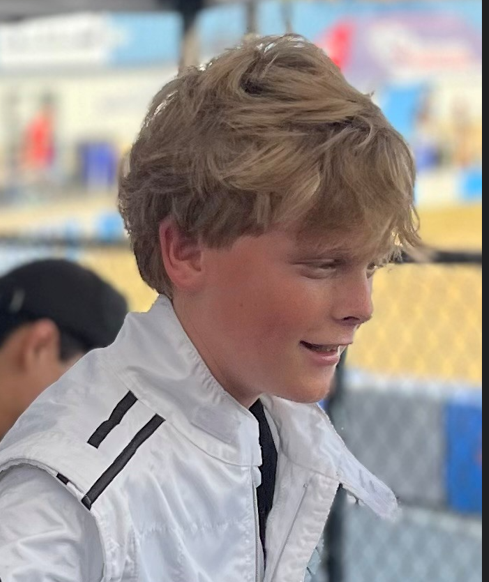 Blond-haired person in a white jacket smiling. Outdoors, blurry background.