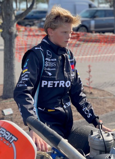 Young person in black racing suit, standing by kart, outdoors.