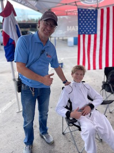Man and boy posing with thumbs up next to a US flag. They are outdoors under a tent.
