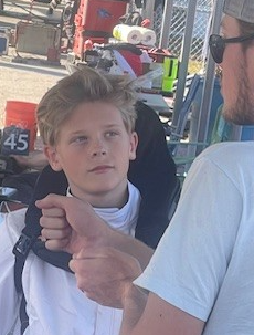 Young person in fencing gear, looking up, with a person adjusting their equipment outdoors.