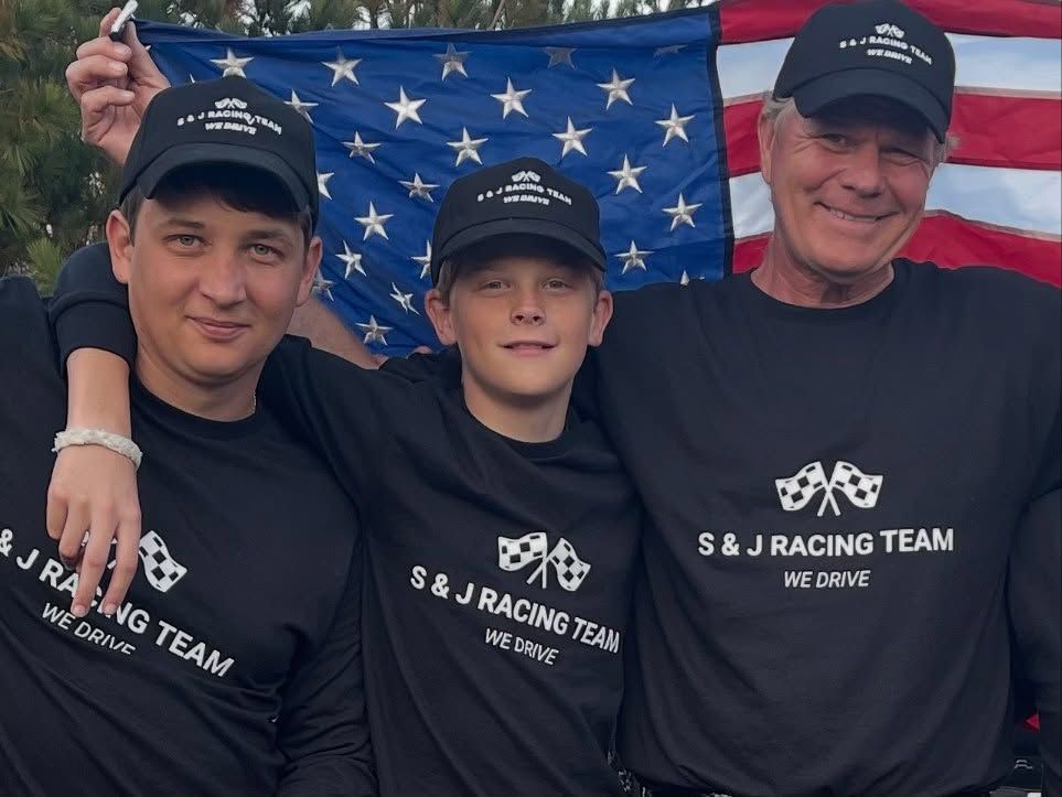 Three people in matching black shirts pose in front of an American flag.