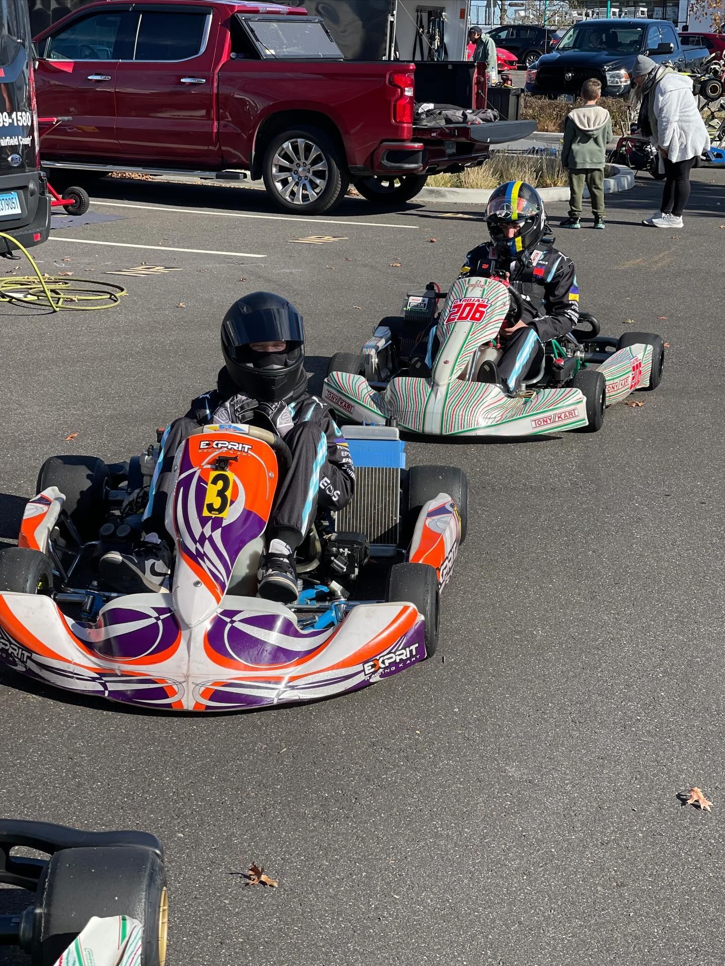 Two go-karts on asphalt track, drivers in helmets, colorful vehicles, onlookers in background.
