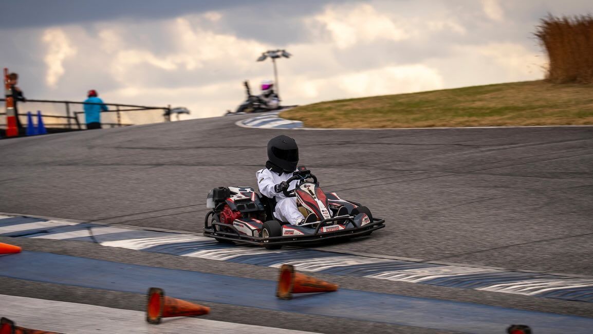 Go-kart driver in helmet and suit racing around a track, orange cones lining the edge.