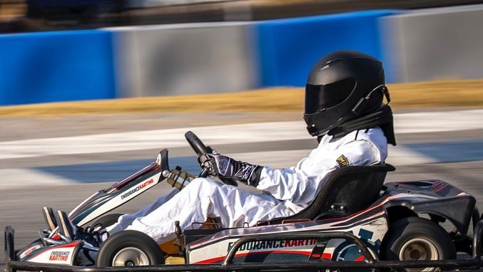 Go-kart driver wearing a black helmet and white racing suit on a track with blue and white barriers.
