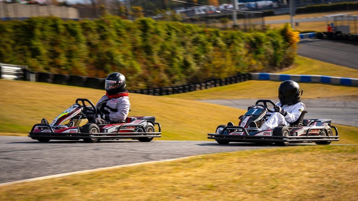 Two go-karts racing on a track, blurred background of trees and structures.