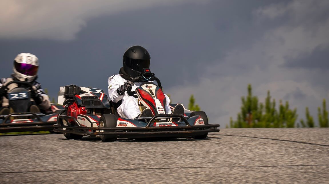 Two go-karts racing on a track, one in front. Dark helmets and suits. Cloudy sky backdrop.