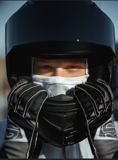 Person in racing gear adjusts helmet, wearing gloves and a face covering.