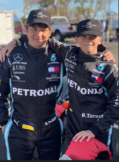 Two people in black Petronas racing suits, one with an arm around the other's shoulder, smiling outdoors.