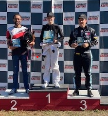 Three people on a podium at a kart racing event. One stands at first, others on second and third. All hold trophies.