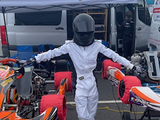 Person in a white racing suit and helmet stands between two go-karts with arms outstretched.