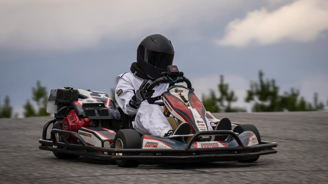 Go-kart driver in a helmet and racing suit on a track, with a cloudy sky in the background.