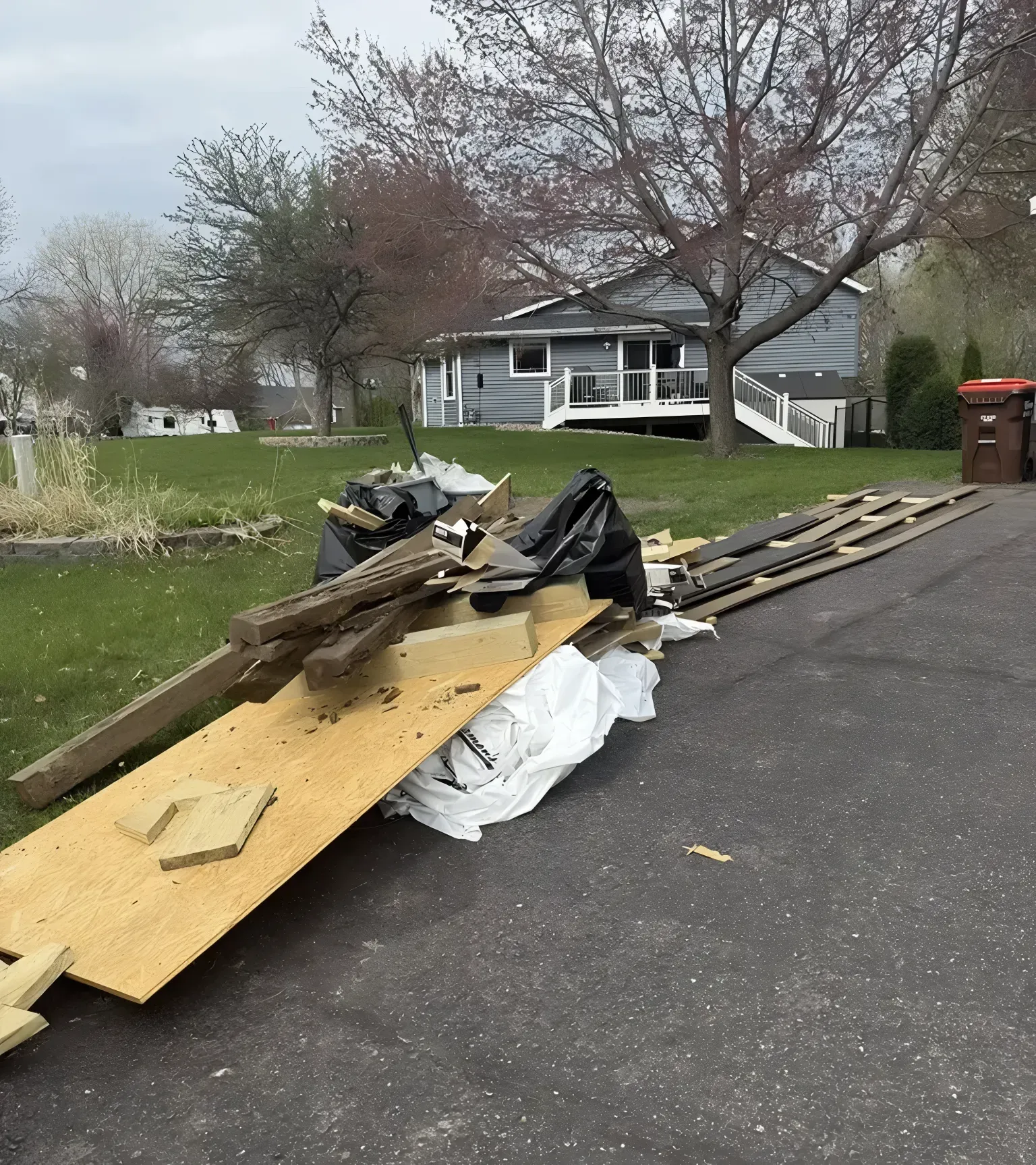 Pile of construction debris on a driveway in front of a gray house with a porch; overcast day.