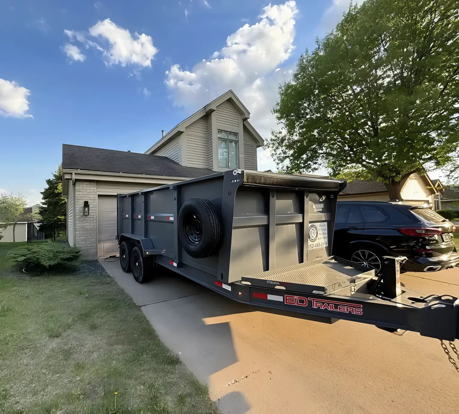 A gray dump trailer is parked on a driveway in front of a two-story house and a black SUV.