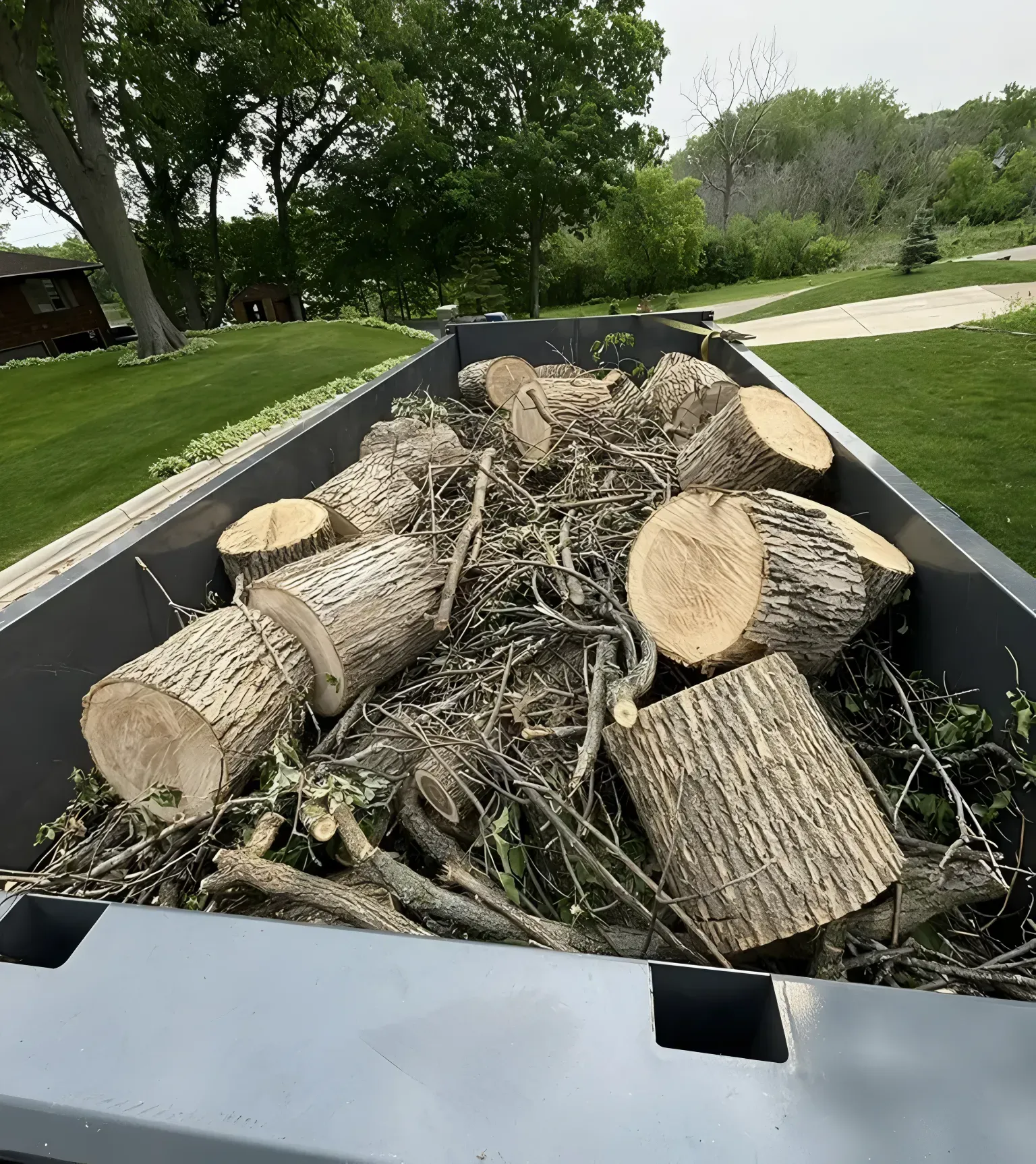 A gray truck bed filled with cut logs and branches, surrounded by green grass and trees.
