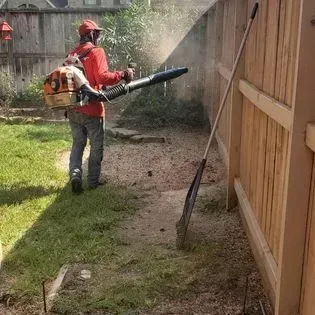 A man is using a leaf blower in a backyard next to a wooden fence.