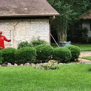 A man is standing in front of a brick building cutting bushes in a yard.