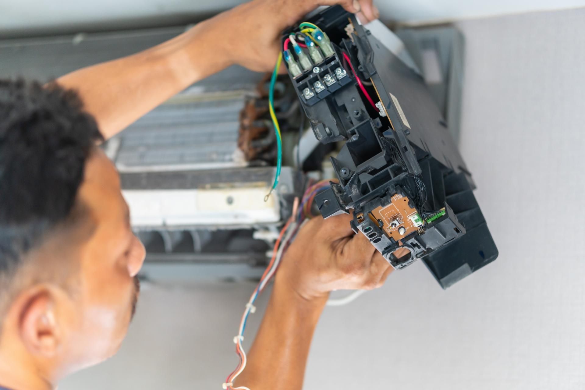 A technician works on the open electrical control panel of a wall-mounted air conditioner.
