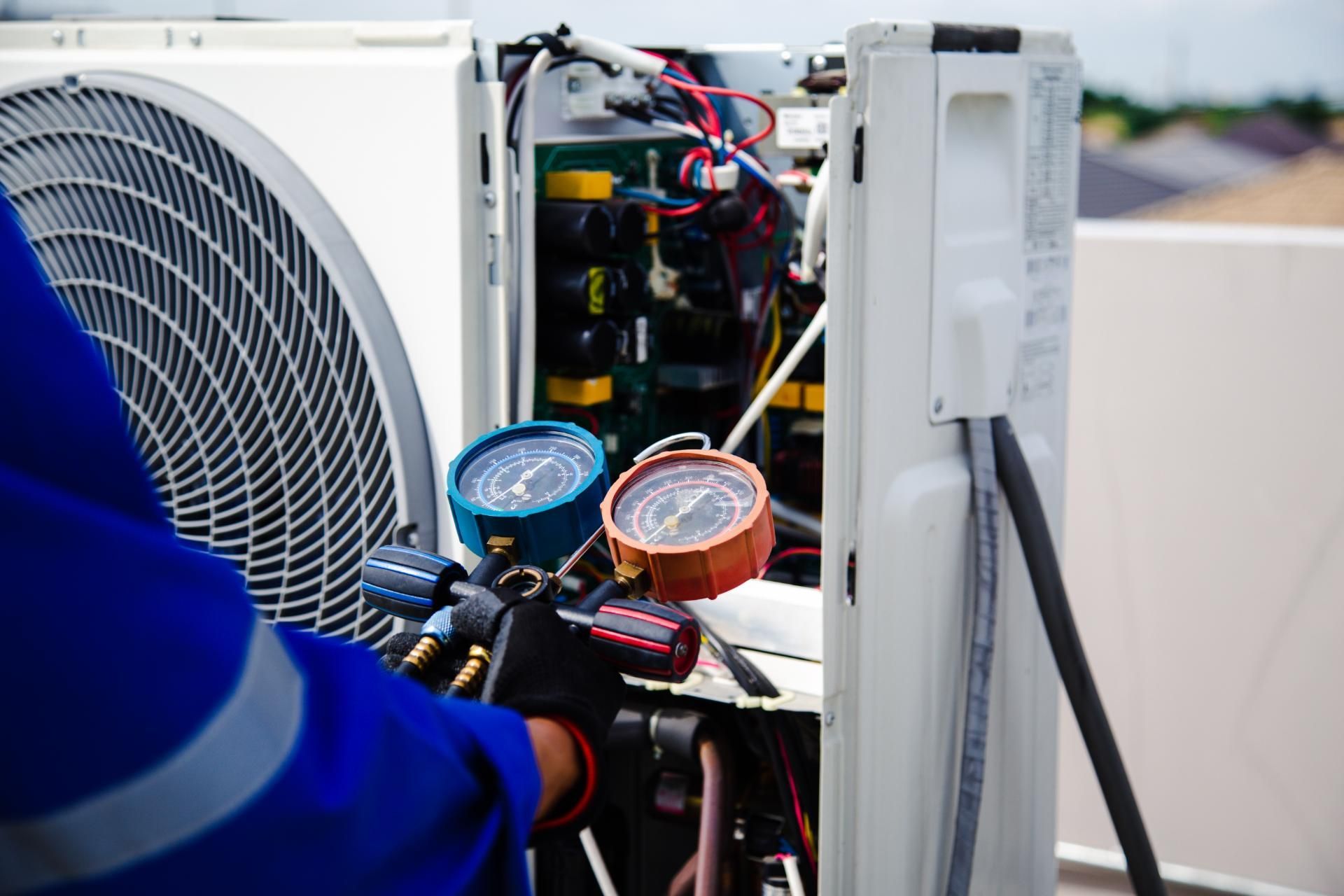 A technician in a blue uniform checks the pressure gauges on an outdoor air conditioning unit's open electrical panel.