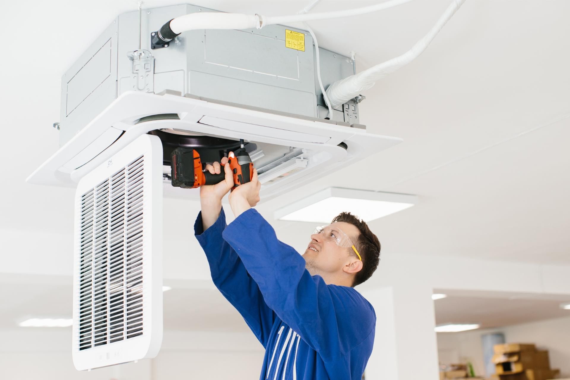 Technician in blue uniform installing a ceiling-mounted HVAC unit with a power drill.