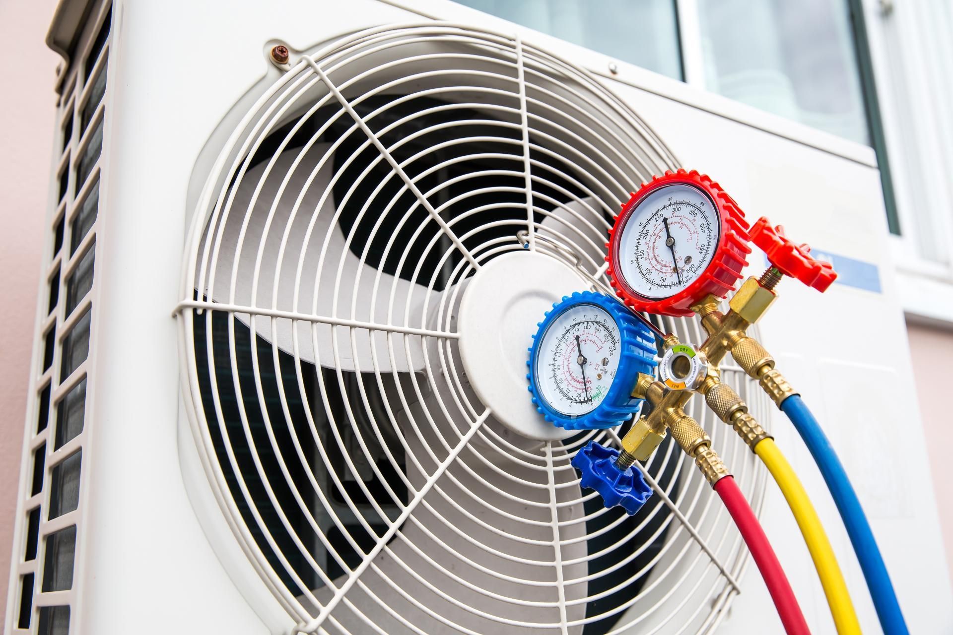 A manifold gauge set with red, blue, and yellow hoses attached to the grill of an outdoor air conditioning unit.