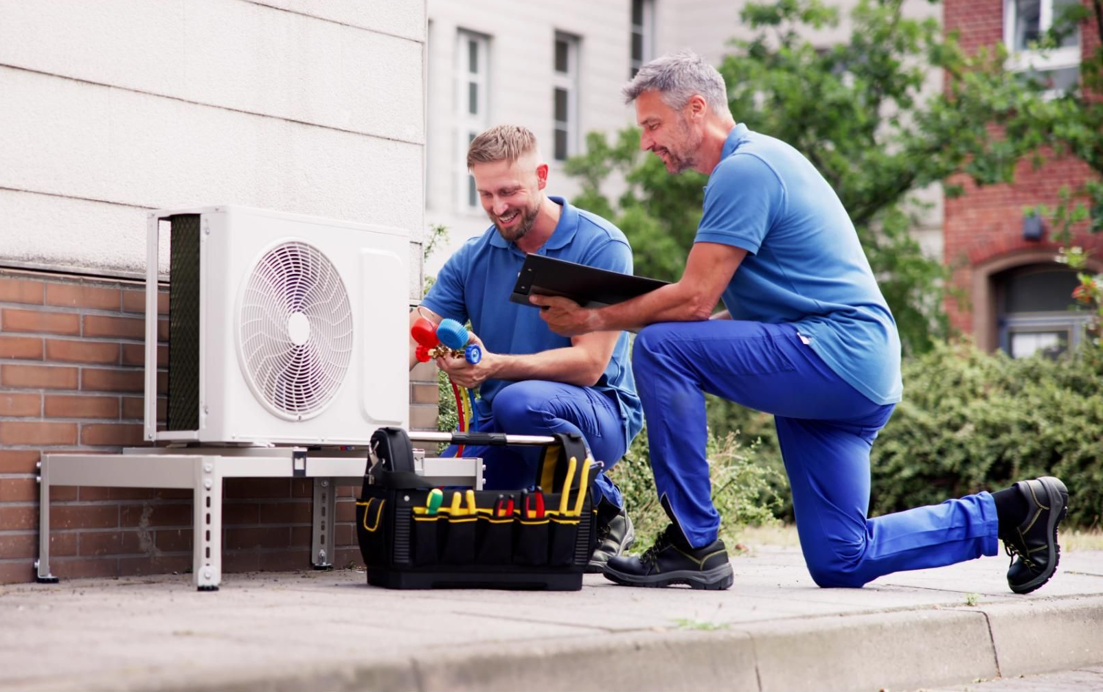 Two technicians in blue uniforms kneeling and working on an outdoor air conditioning unit with tools and a clipboard.