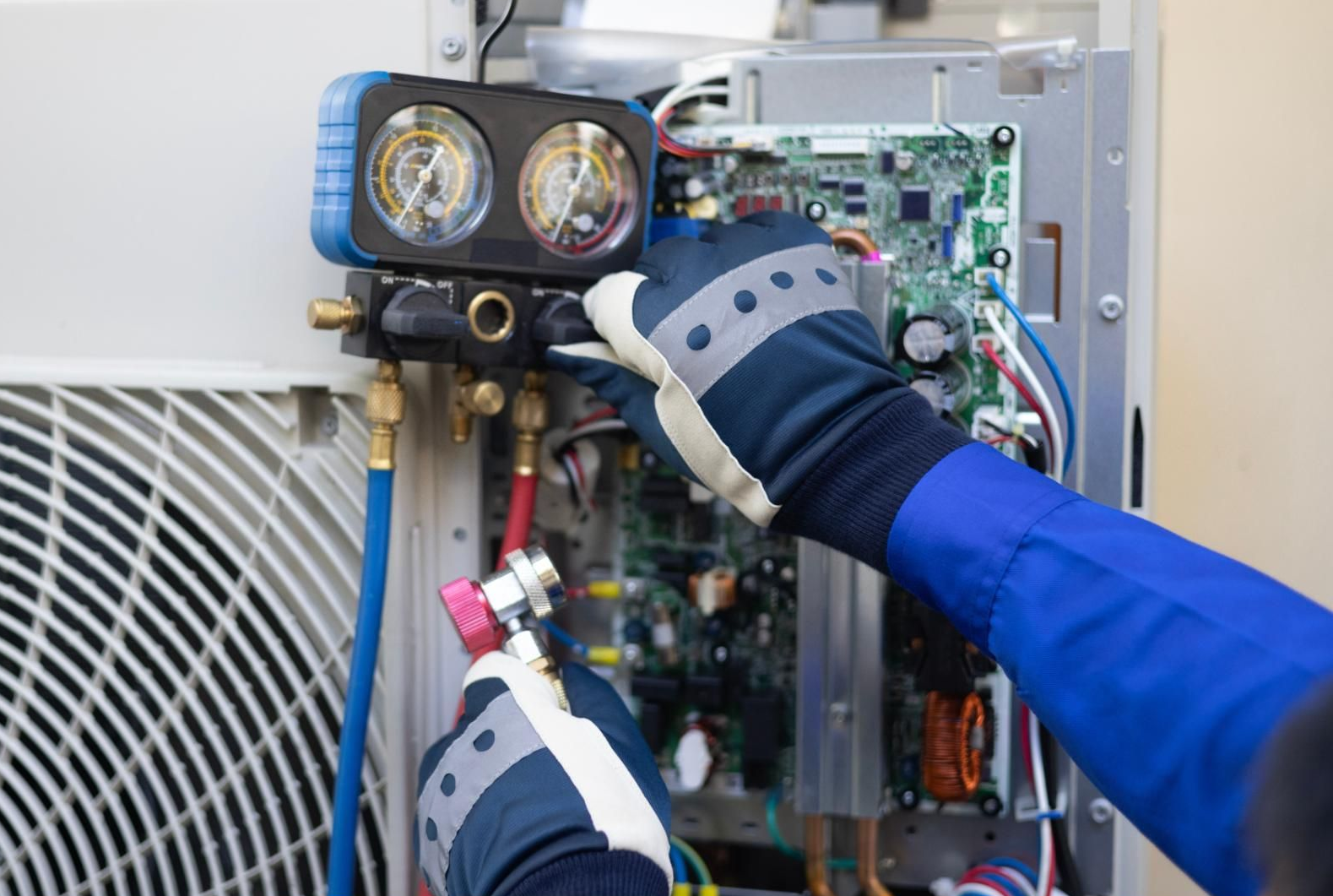 A technician in blue coveralls and gloves uses a manifold gauge set to service an outdoor air conditioning unit.