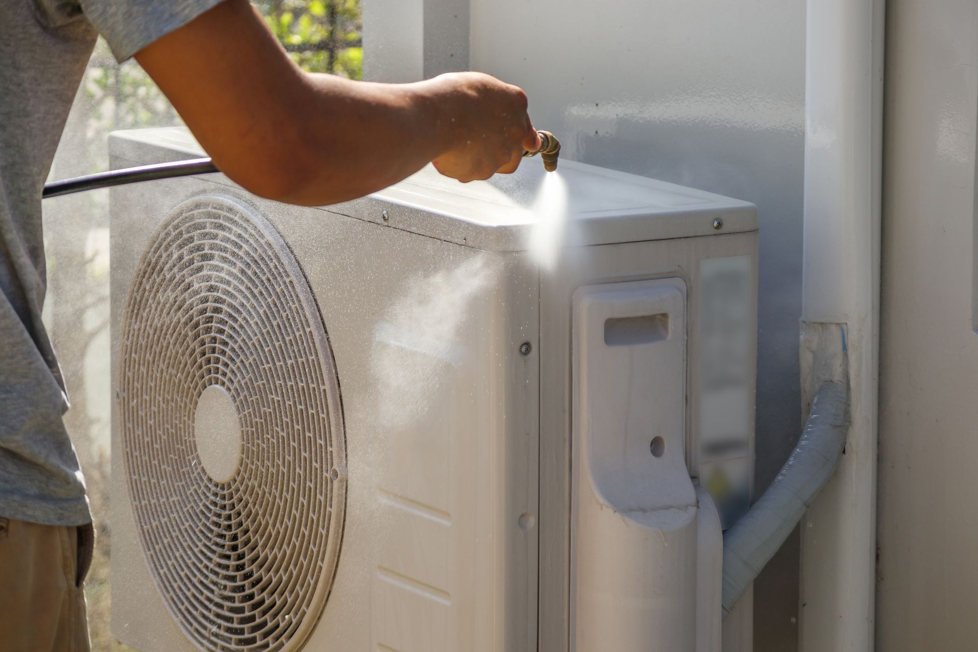 A technician in a red uniform and gloves kneels on grass, working on a white outdoor heat pump unit near a brick wall.
