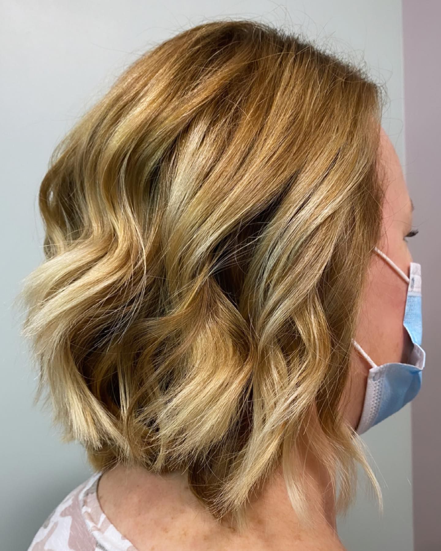 Woman with wavy blonde bob haircut, wearing a surgical mask, against a plain background.