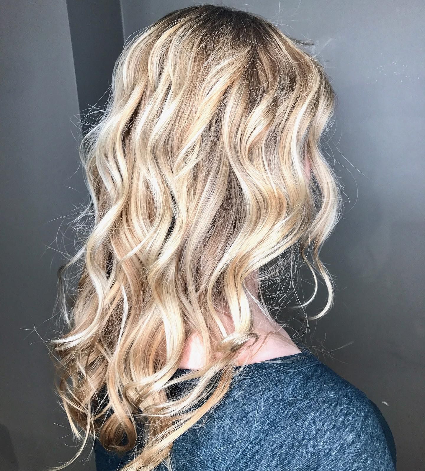 Blonde hair with loose waves, possibly highlighted. A woman's back is visible, wearing a gray shirt.