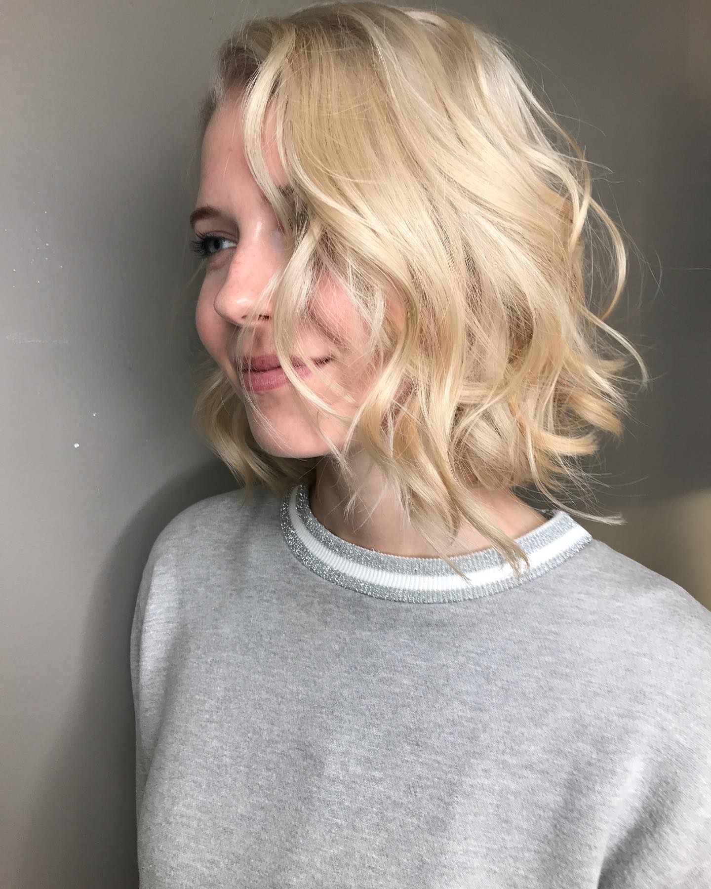 Woman with blonde wavy bob haircut, smiling, wearing a gray sweater.
