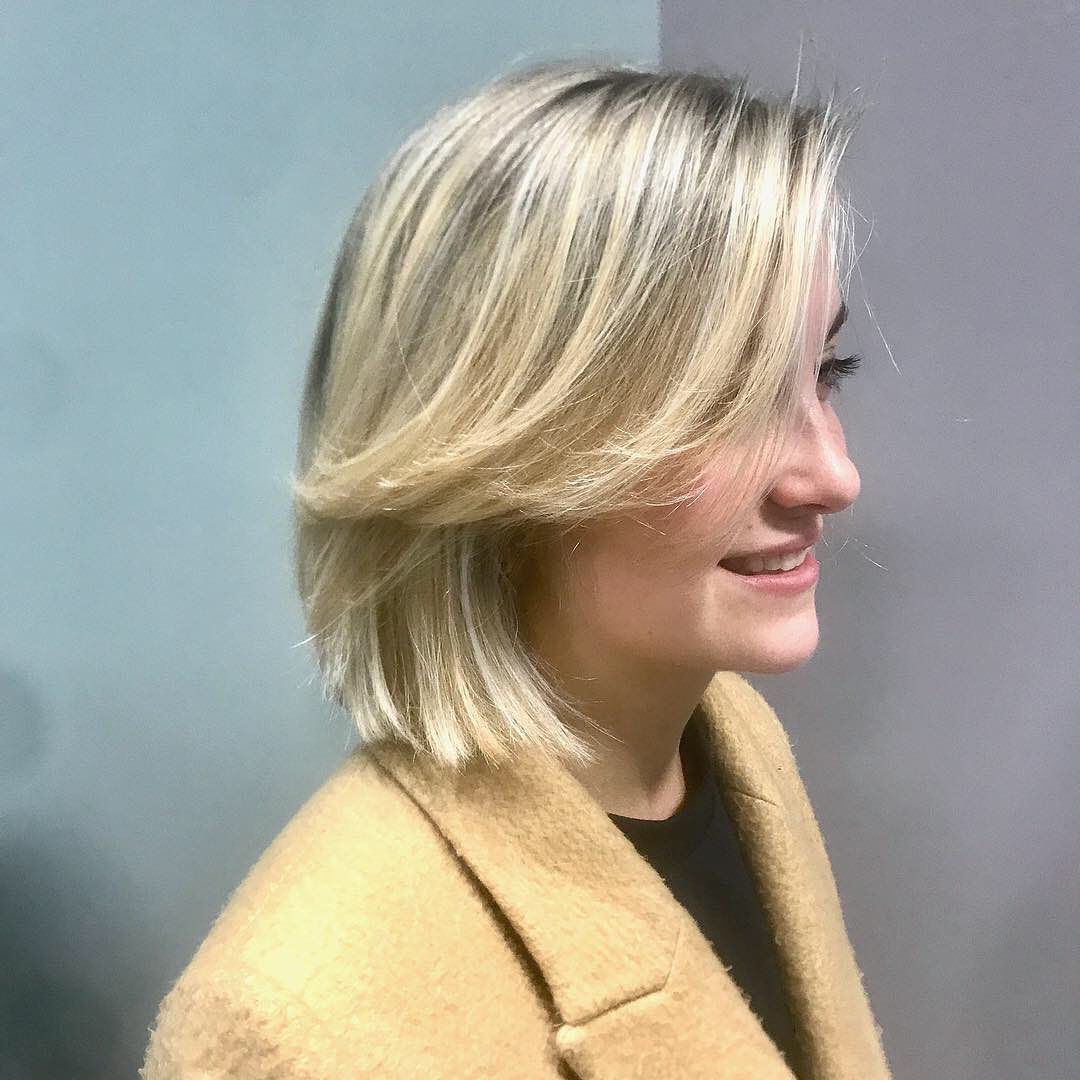 Blonde woman in tan coat smiles, profile view; short layered hair, neutral background.