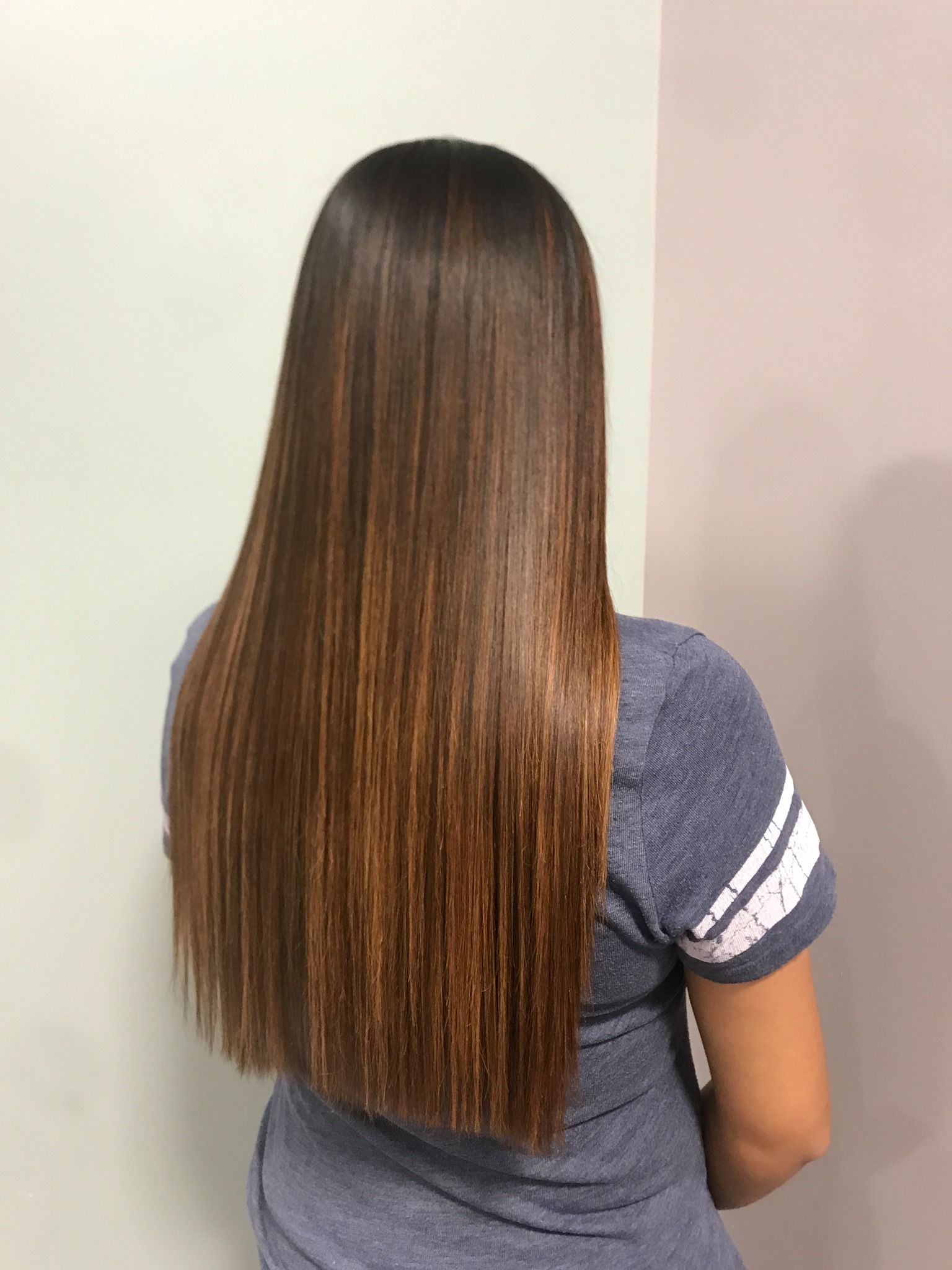 Woman with long, straight, brown hair; back view; against a light-colored wall; wearing a blue t-shirt.