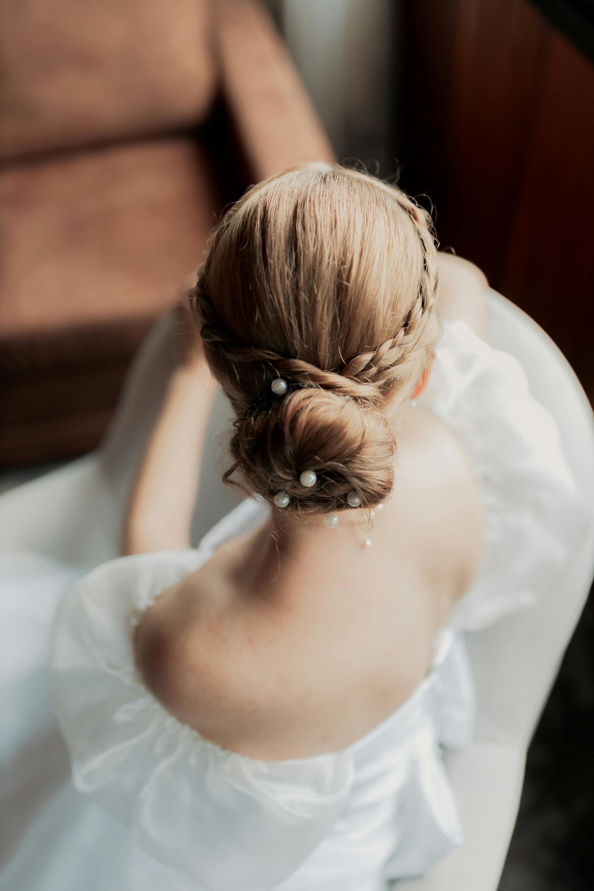 Bride in a white gown, seated with her updo hairstyle visible. Pearls adorn the bun.