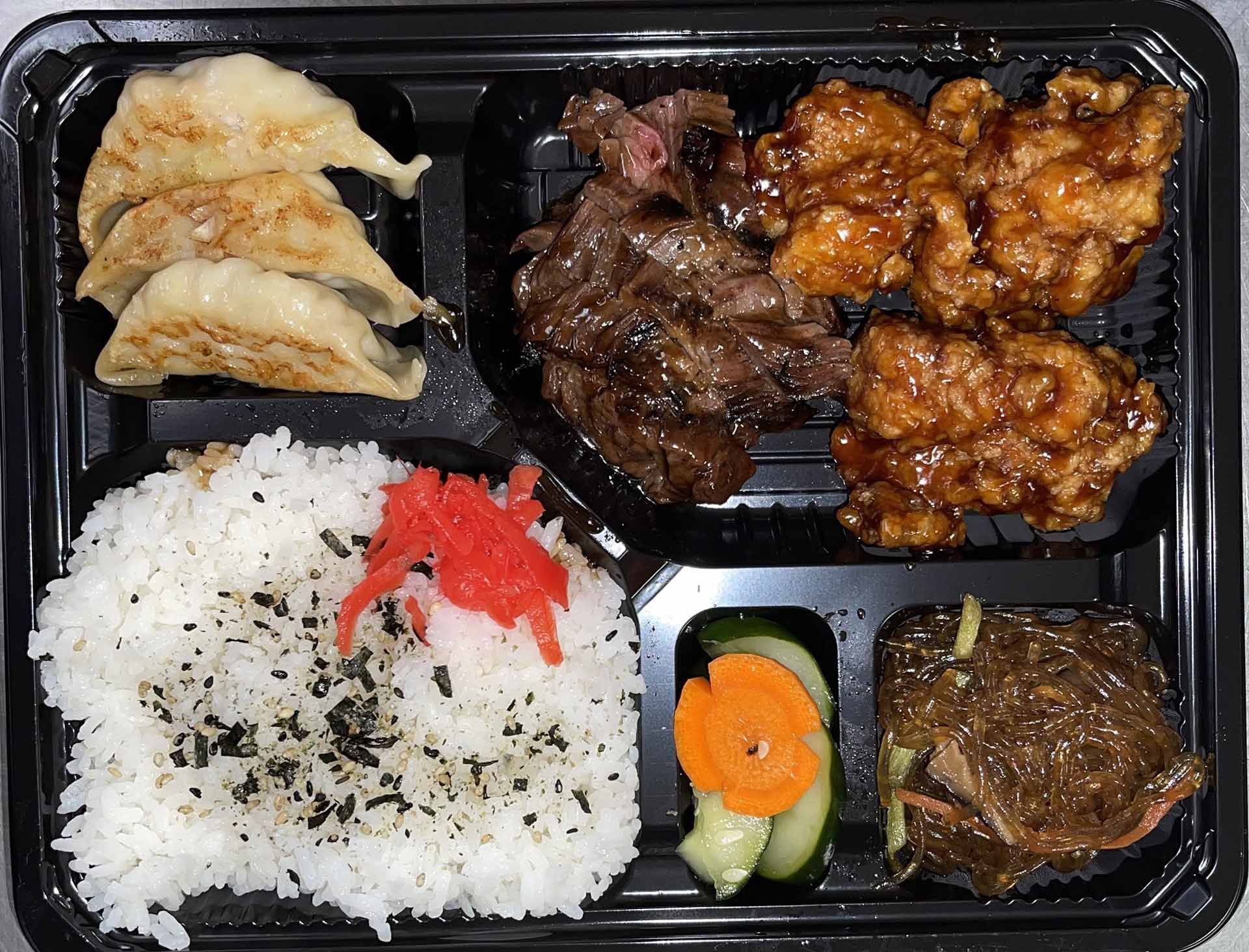 Bento box with gyoza, steak, chicken, rice with nori, pickled ginger and vegetables