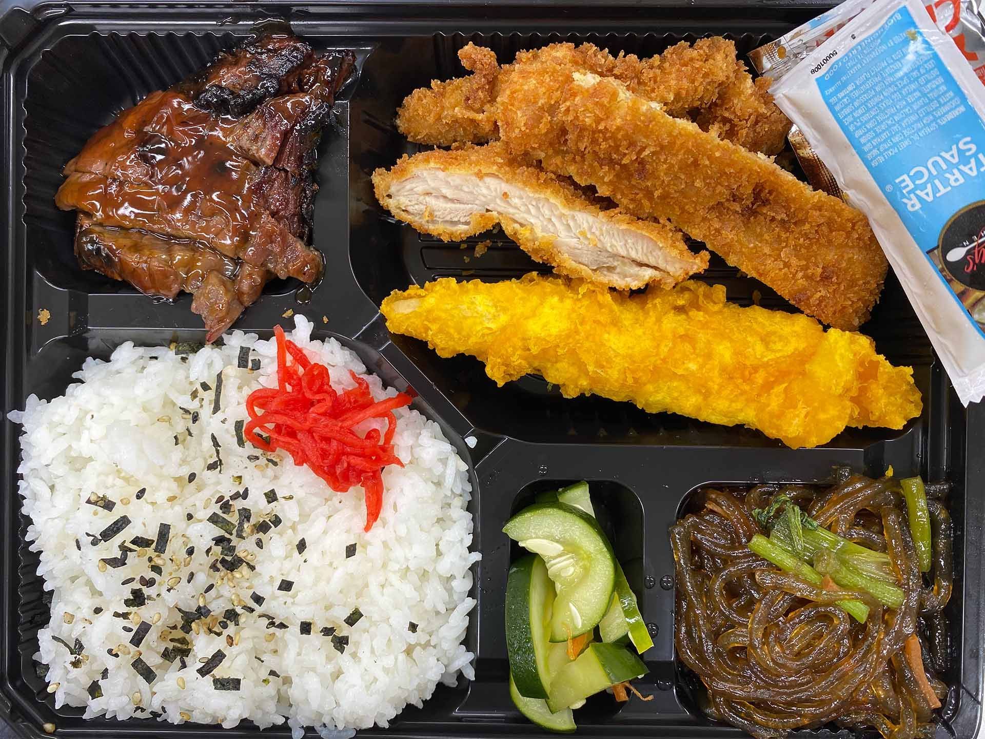 Bento box with rice, glazed meat, fried chicken, tempura, pickled cucumbers, glass noodles, and tartar sauce packet