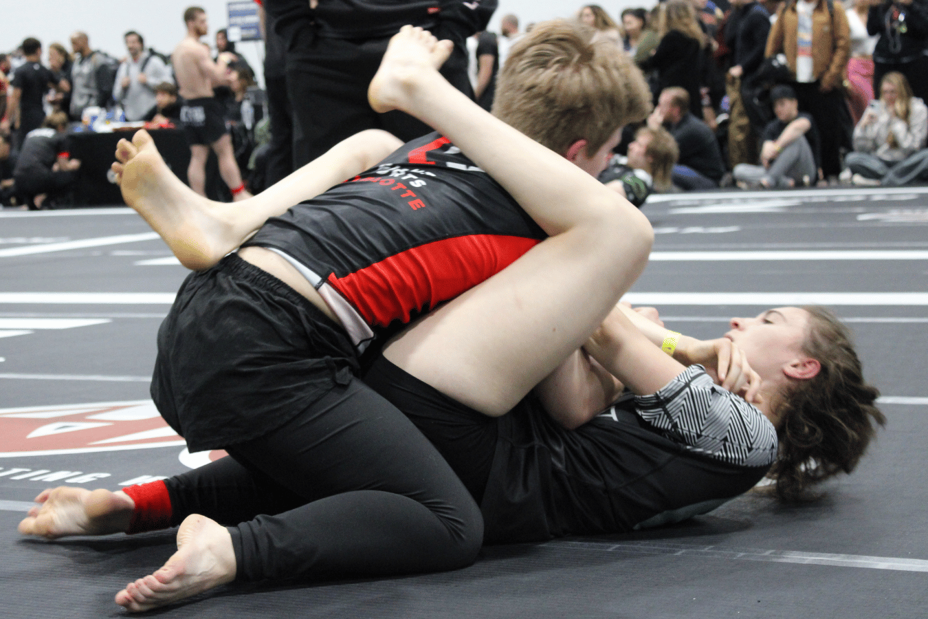 Two kids competing in a Brazilian Jiu-Jitsu match, one applying a top control position on gray mats.