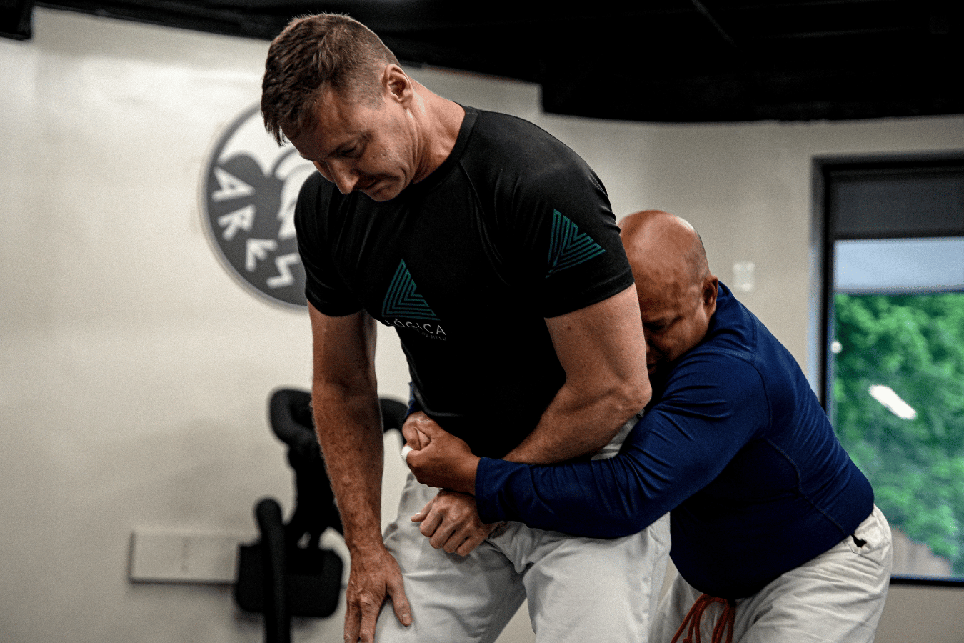 Two adults practicing a standing self-defense technique indoors.