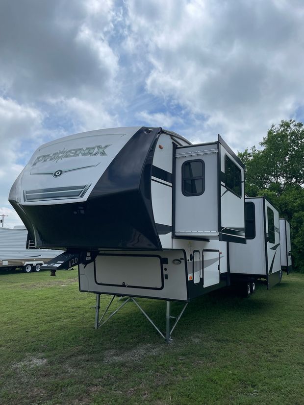 A white and black Phoenix fifth-wheel RV parked on green grass under a cloudy sky.
