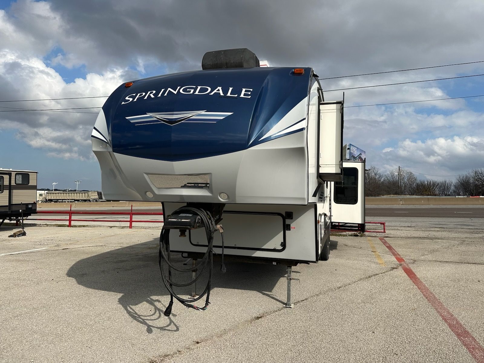 Blue and white Springdale fifth wheel camper on a gravel lot with a cloudy sky.