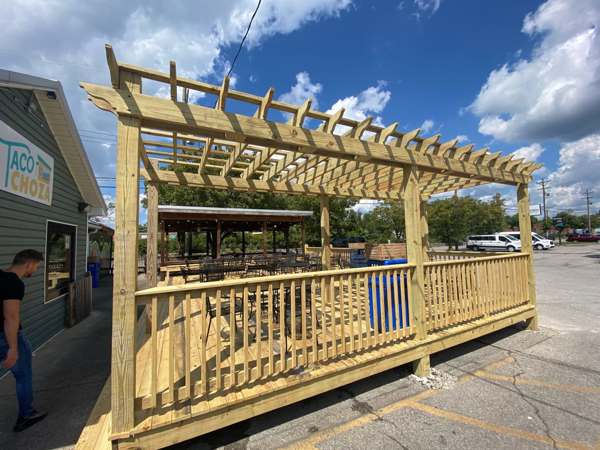 A man is standing next to a wooden pergola in a parking lot.