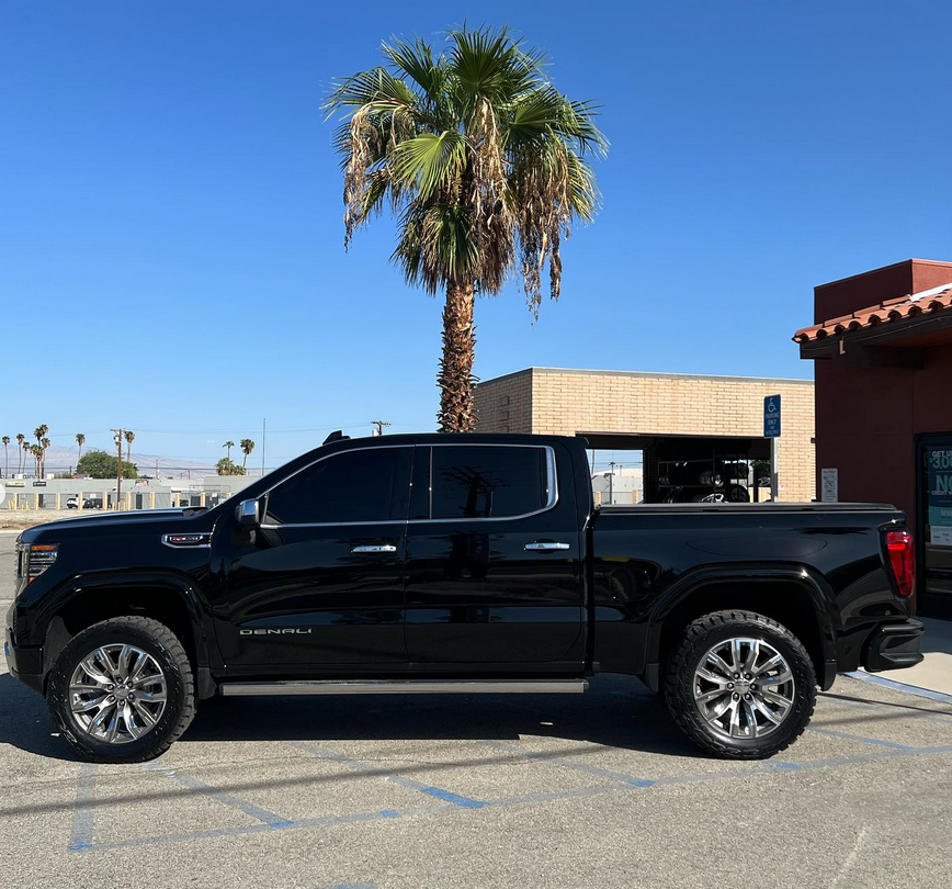 A black truck is parked in a parking lot next to a palm tree.