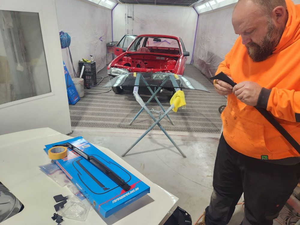 A Man in an Orange Hoodie is Working on a Red Car in a Paint Booth — MJR Auto Glass In Southern Highlands, NSW