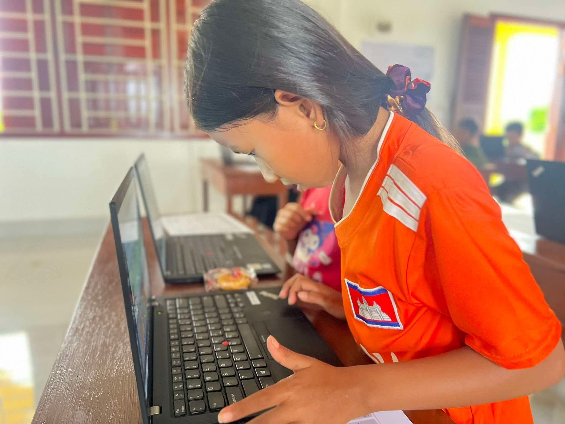 Girl in orange shirt uses a laptop in a classroom; other students are in the background.