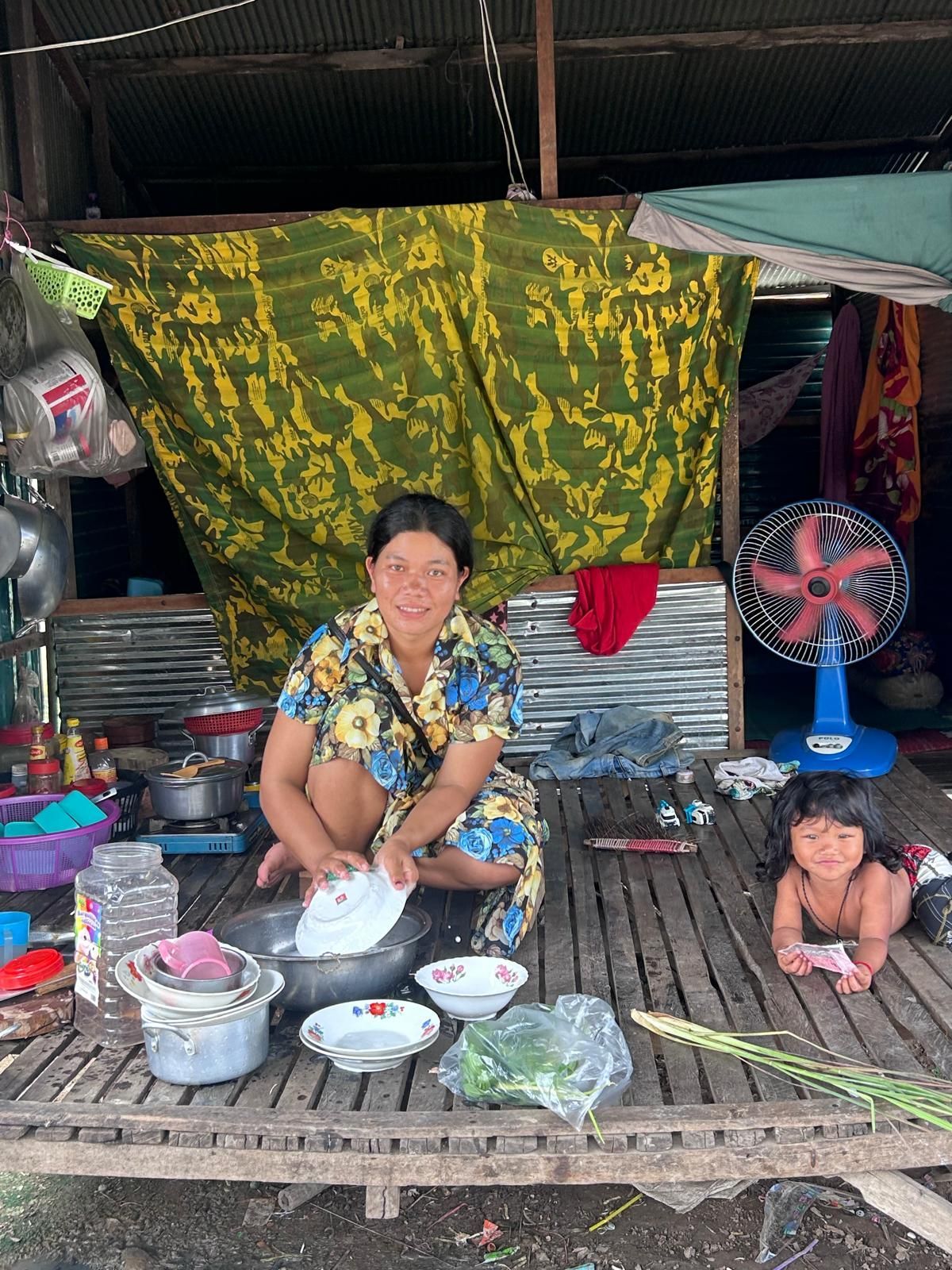 Woman washing dishes outside her home. Child looks on. Tropical setting.