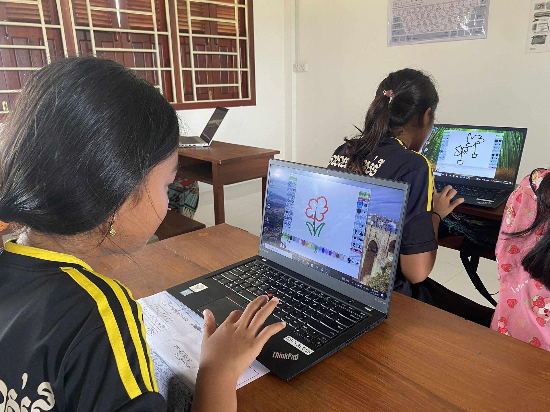 Children in a classroom use laptops, looking at a projector screen. Wooden desks and windows are visible.