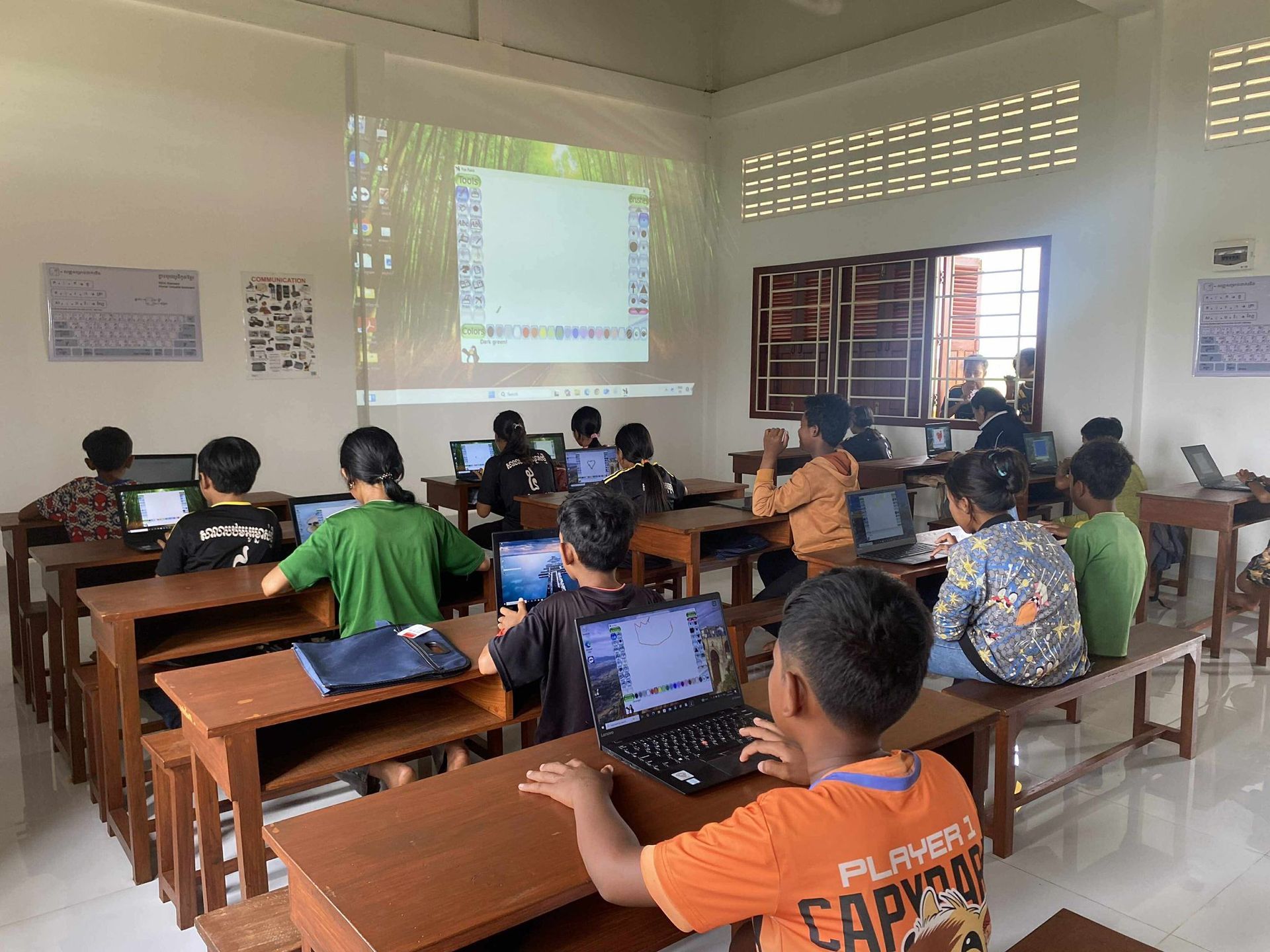Children in a classroom with laptops, looking at a projected screen. Wooden desks, sunny room.