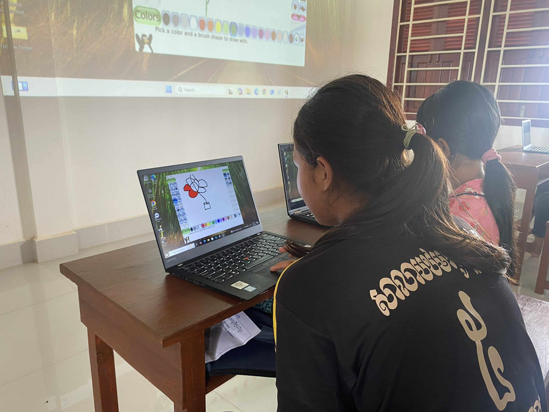Students using laptops, one with a drawing on screen, classroom setting, projector in background.