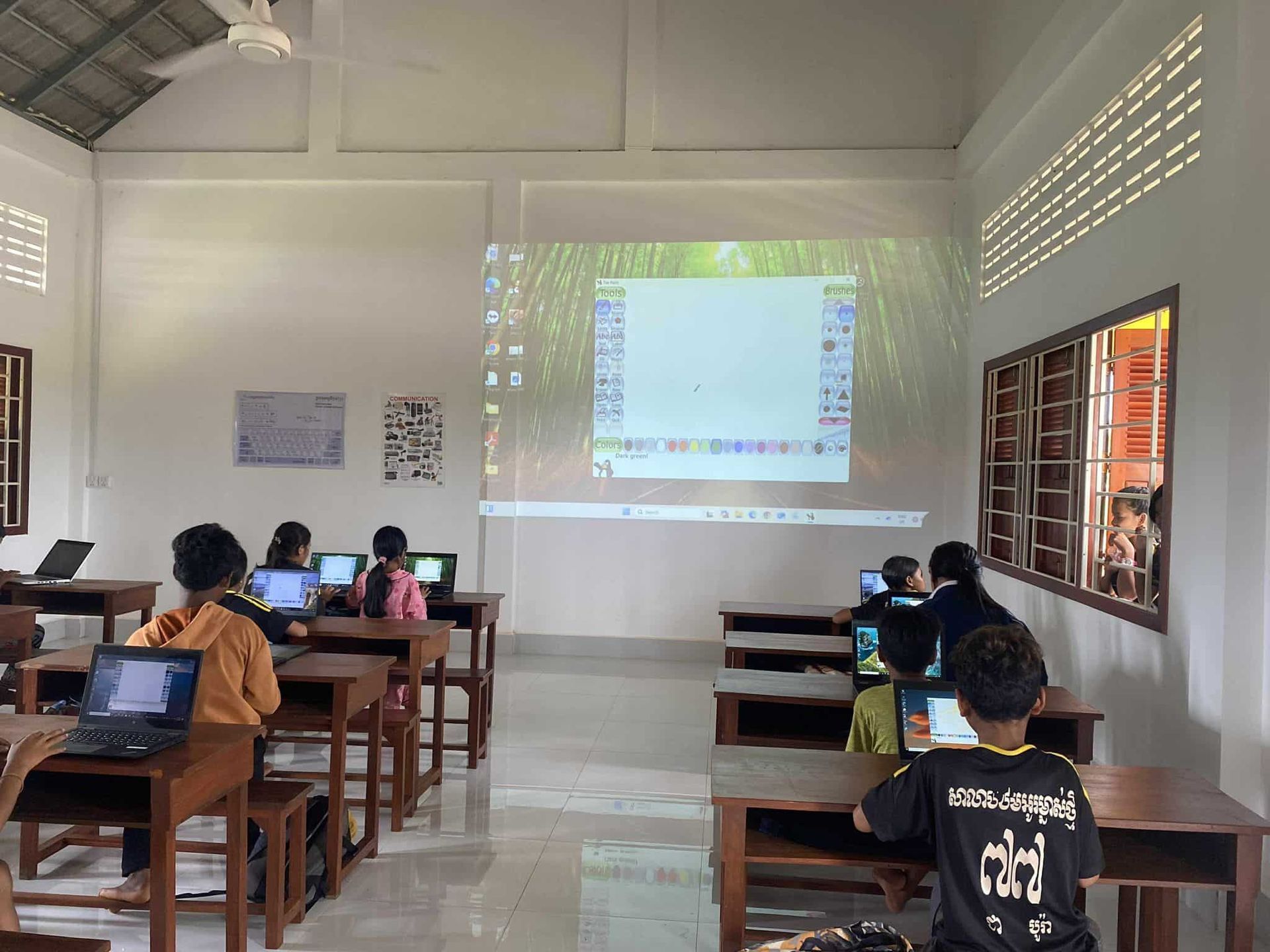 Students at desks in a classroom, focused on laptops, projected screen up front. Windows let in light.