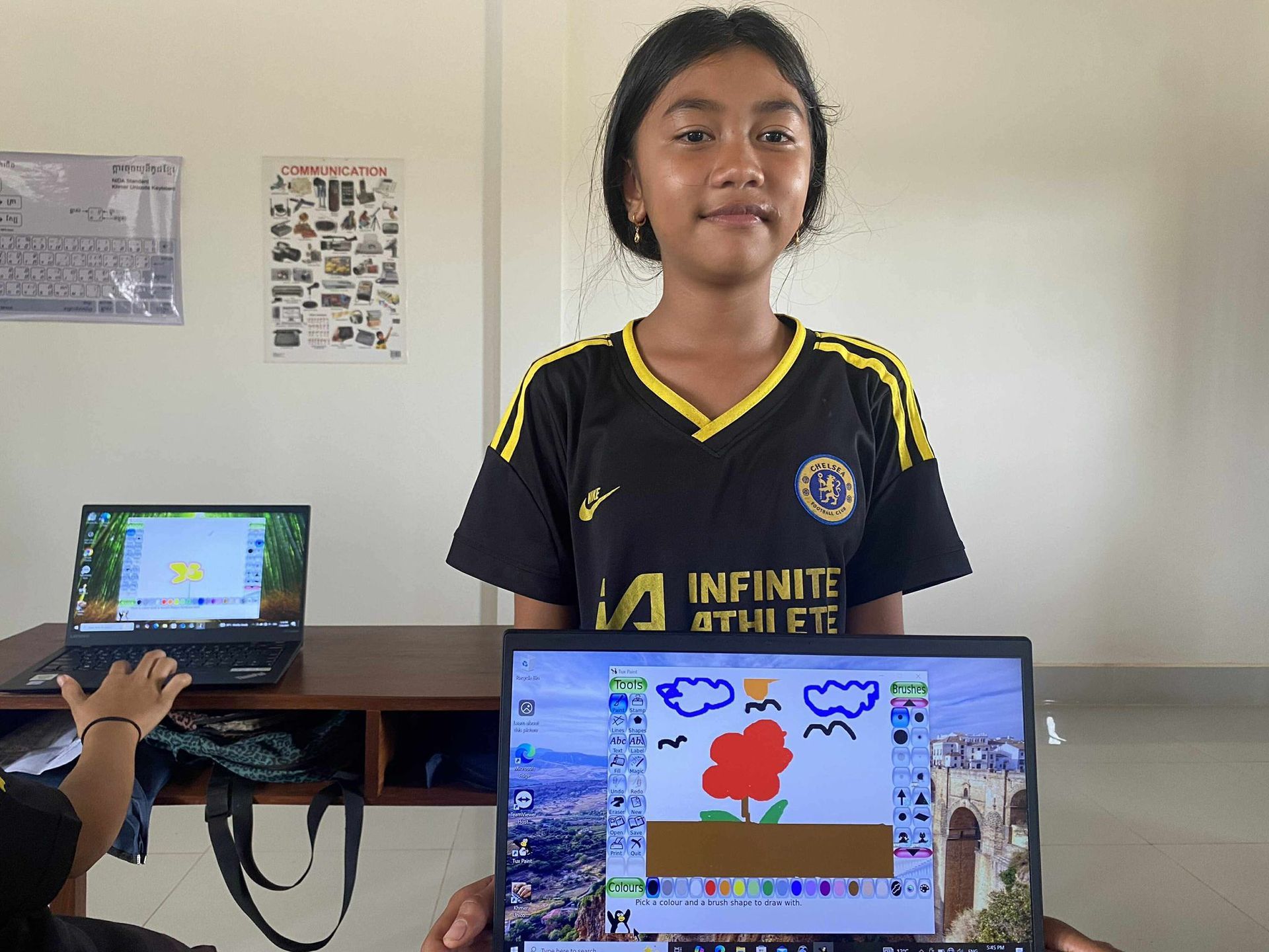 Girl in black shirt holding laptop, displaying a MS Paint drawing of a flower, clouds, and birds. Classroom setting.