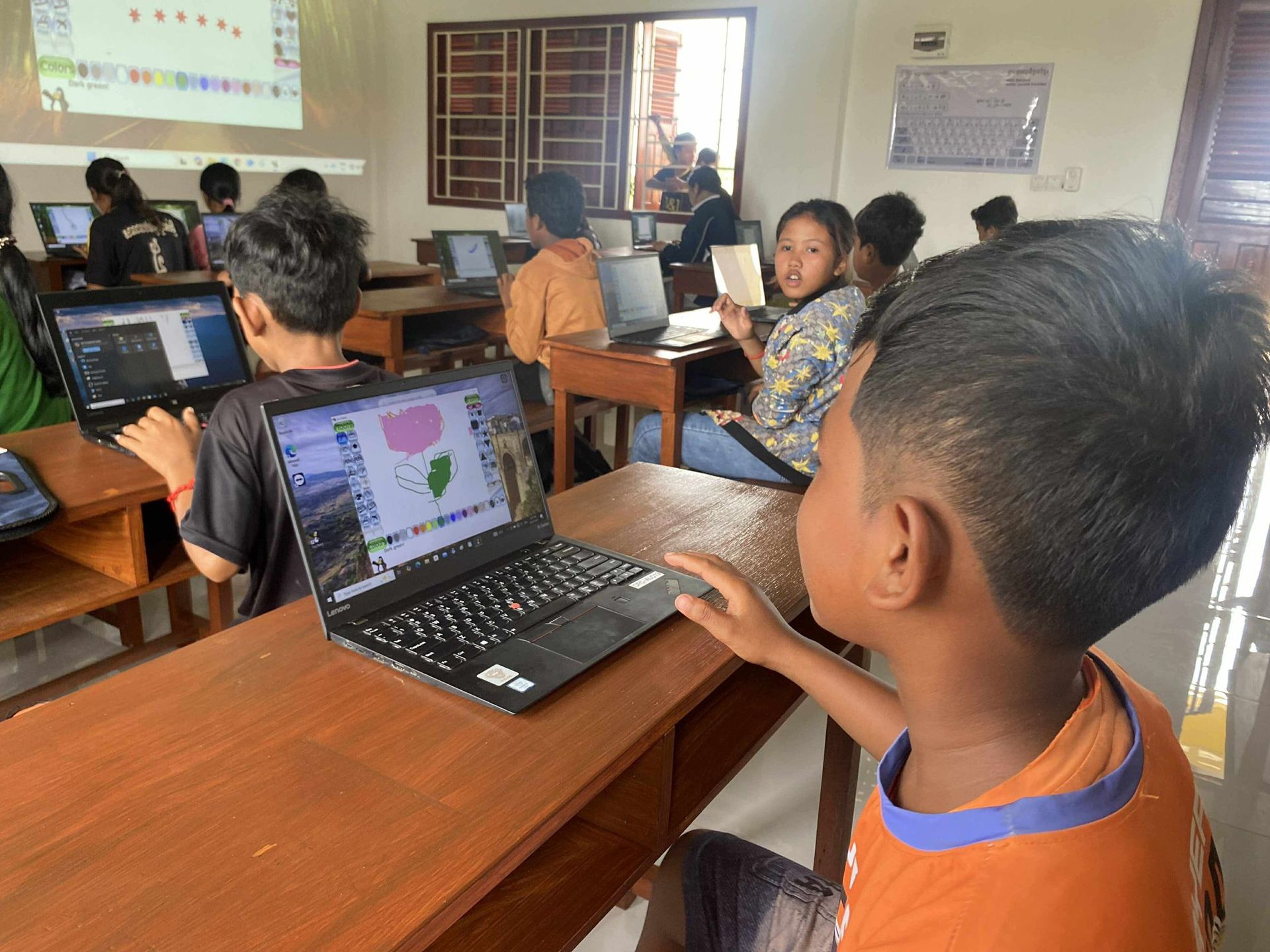 Children in a classroom using laptops; a student looks at screen displaying a drawing; a projector shows a lesson.